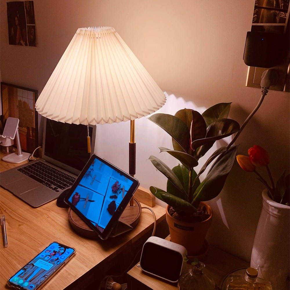 Pleated lamp on a desk with a tablet resting across its base beside a laptop, phone, plant and clock