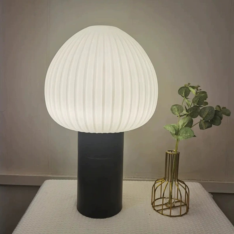 White ribbed-shade lamp with black base on a pale cloth beside a gold-toned wire-outline vase and green leaves