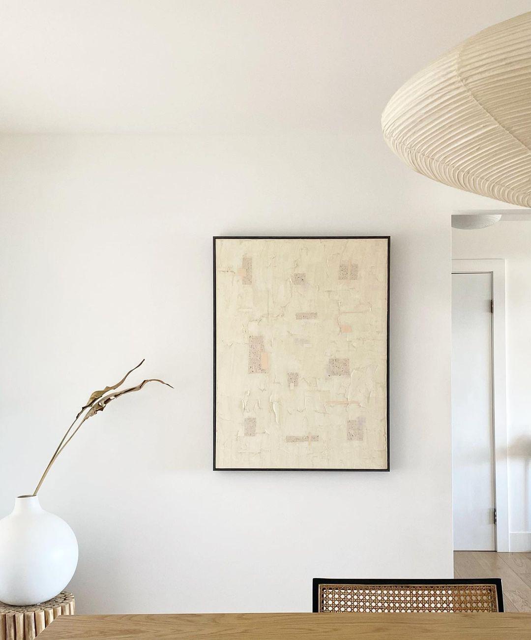 Cropped paper shade above a wood tabletop beside pale textured artwork, a white vase and a cane chair