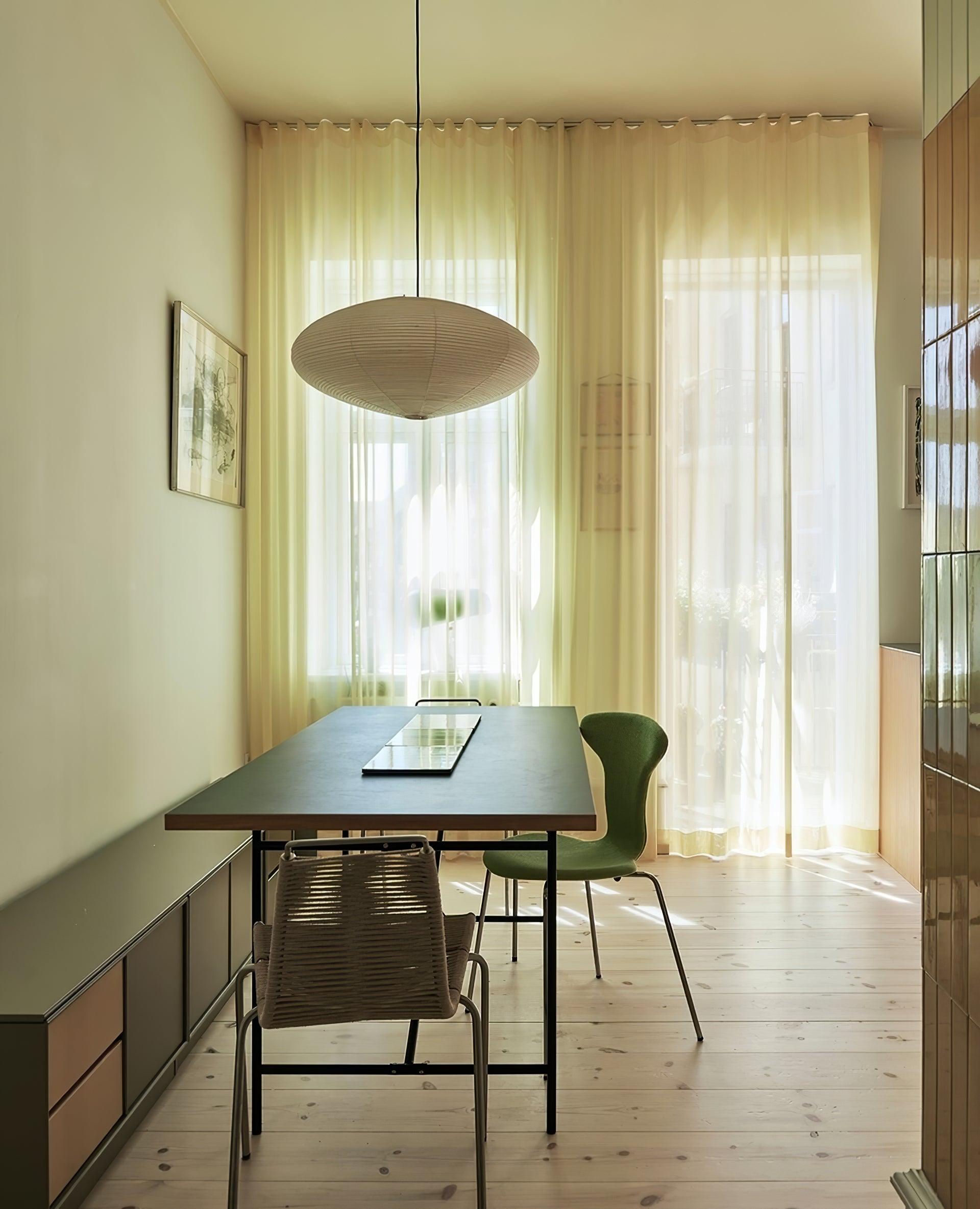Paper pendant above a long dark table with green and woven chairs beside sheer curtains and wood cabinets