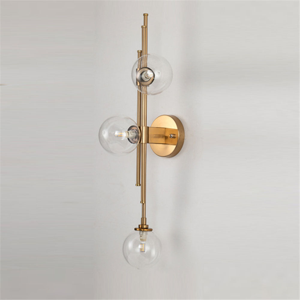 Clear glass globes at three staggered heights on gold-tone rods with a circular wall plate against a pale wall