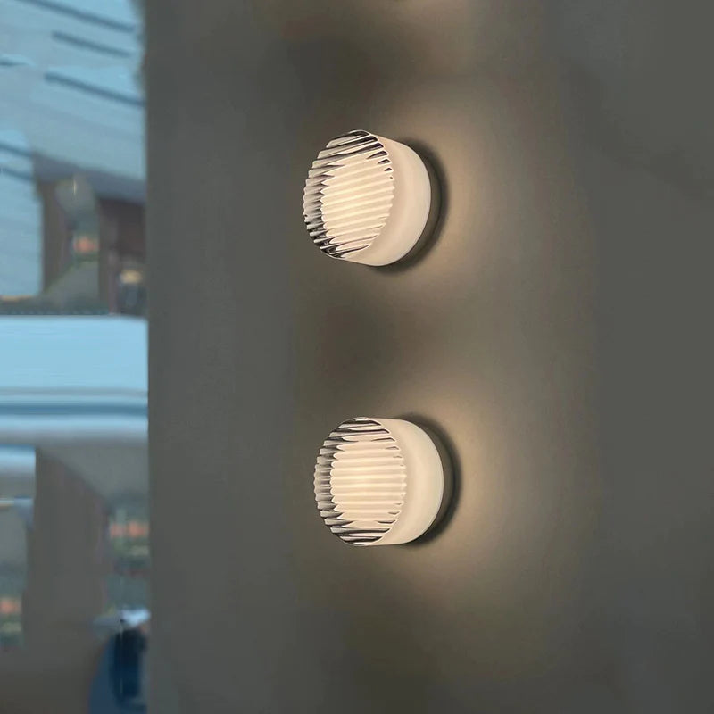 Two white round ribbed lights placed vertically on a grey wall beside a blurred window and shelves