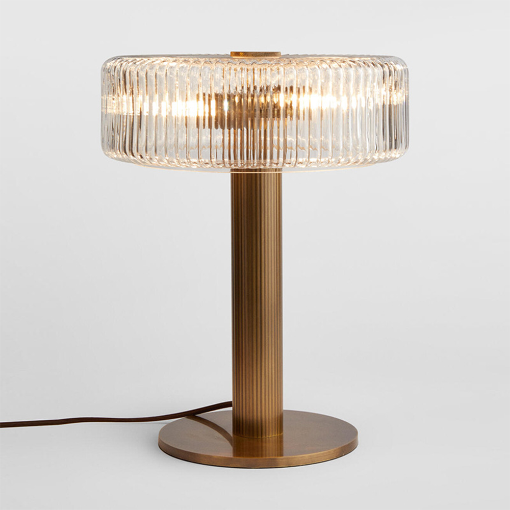 Clear ribbed-glass drum-shade lamp illuminated on a fluted gold-tone column and circular base against pale grey, with a cord to the left