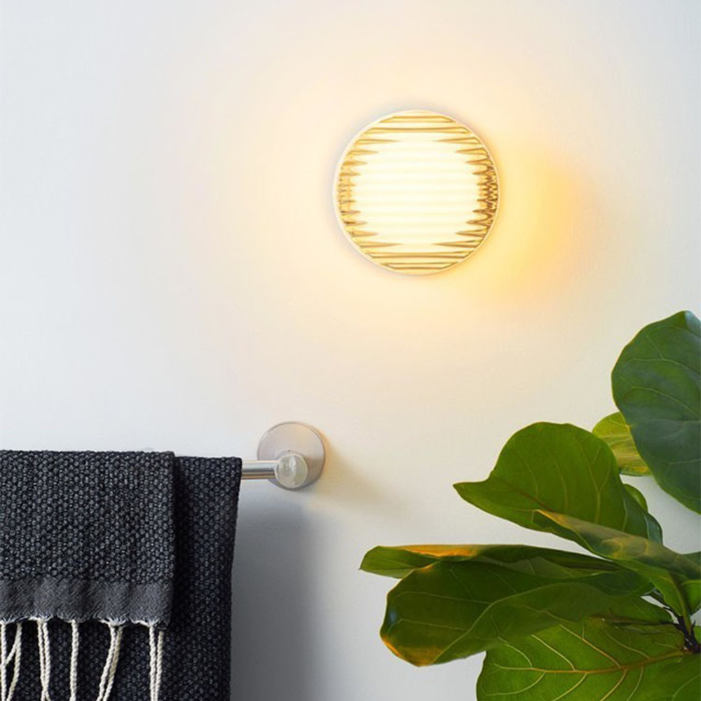 Warm-lit round wall fixture above a dark towel on a rail beside a broad-leaf plant