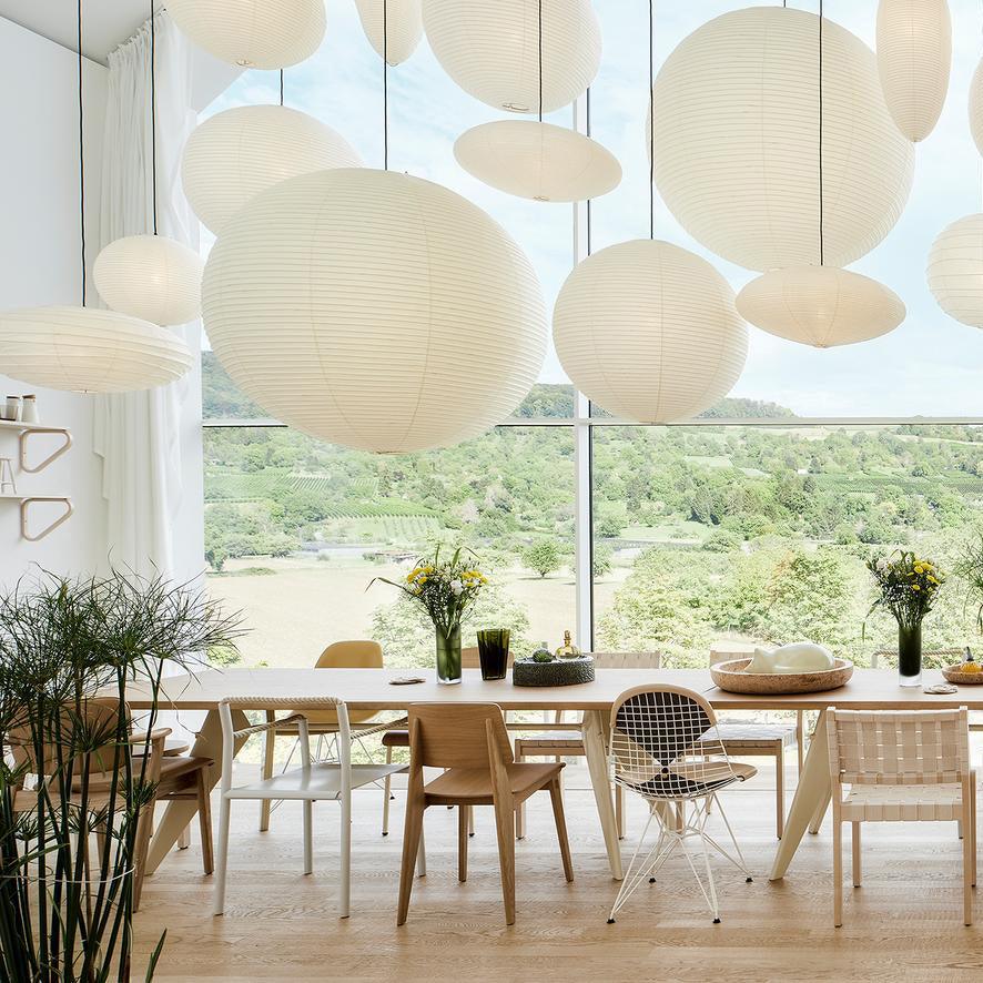 Globe and oval paper shades over a long dining table with mixed chairs and flower vases by a large window
