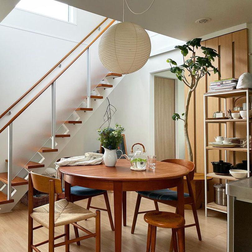 Paper globe above a round wood dining table near an open timber staircase, kitchen shelves and a tree