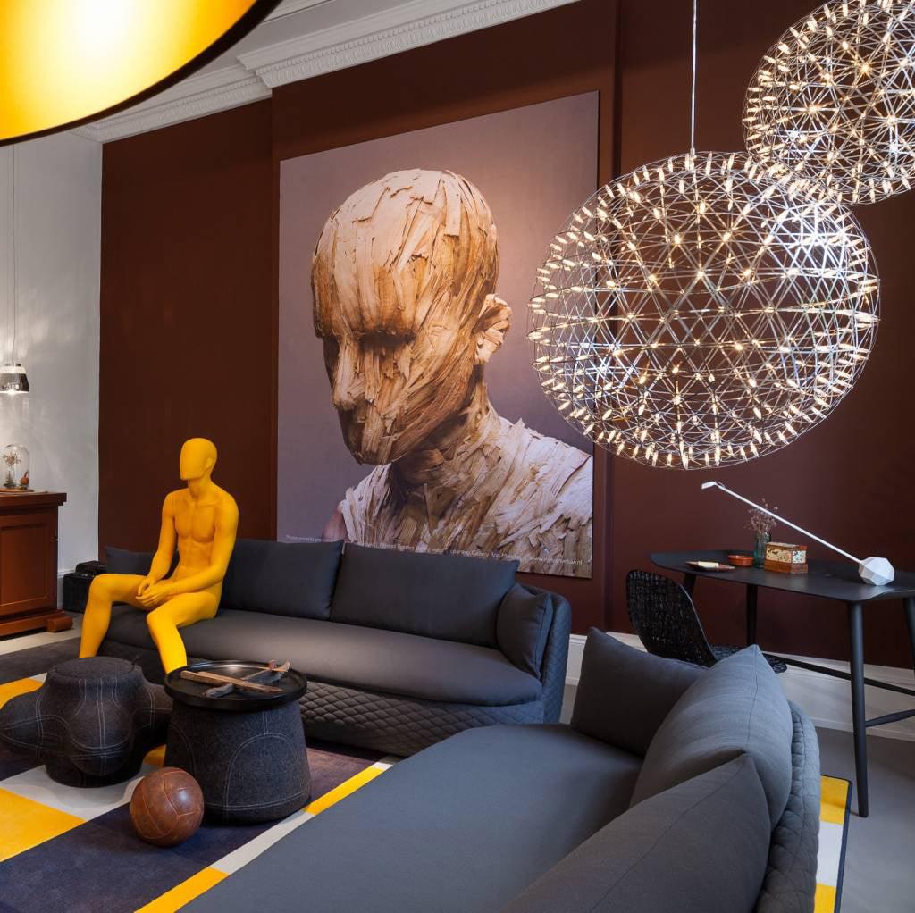 Large glowing globes above grey sofas near a yellow mannequin and sculptural wall art