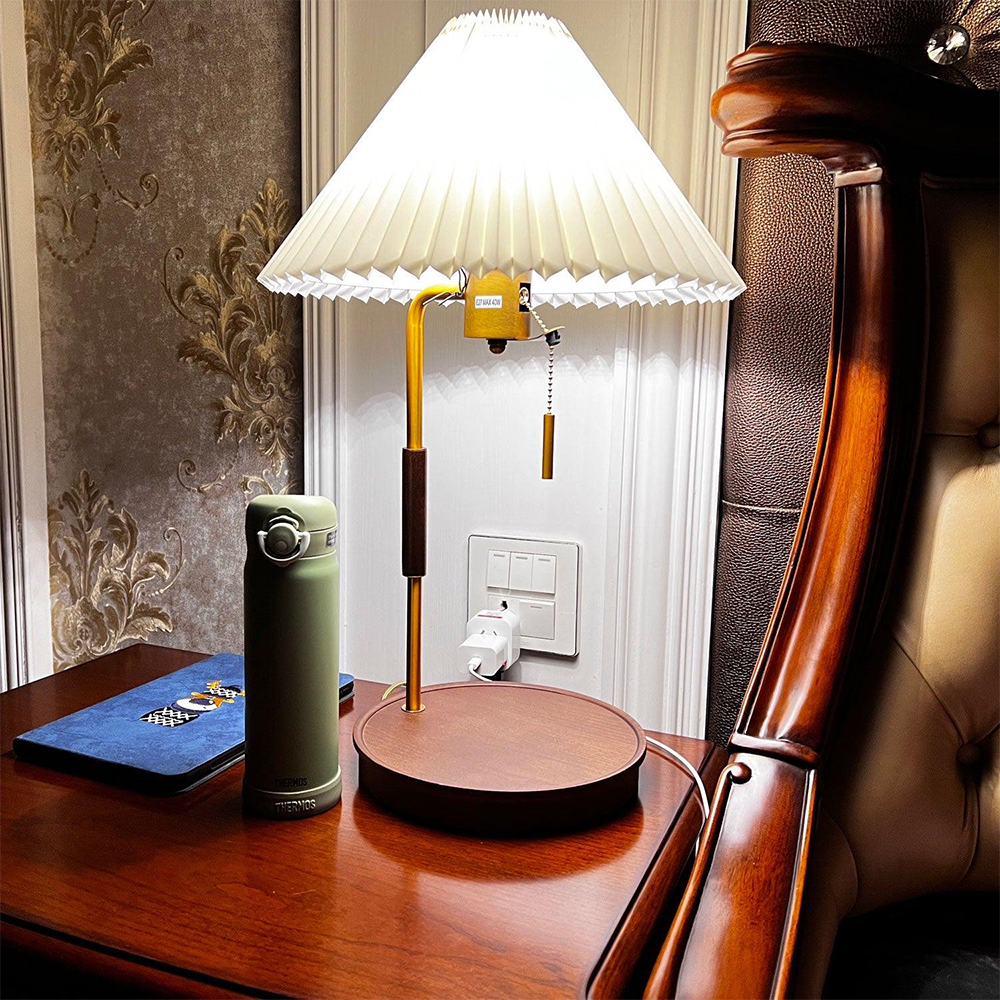 Lit pleated lamp with a brown wood base on a bedside cabinet near a bottle, book and upholstered bedpost