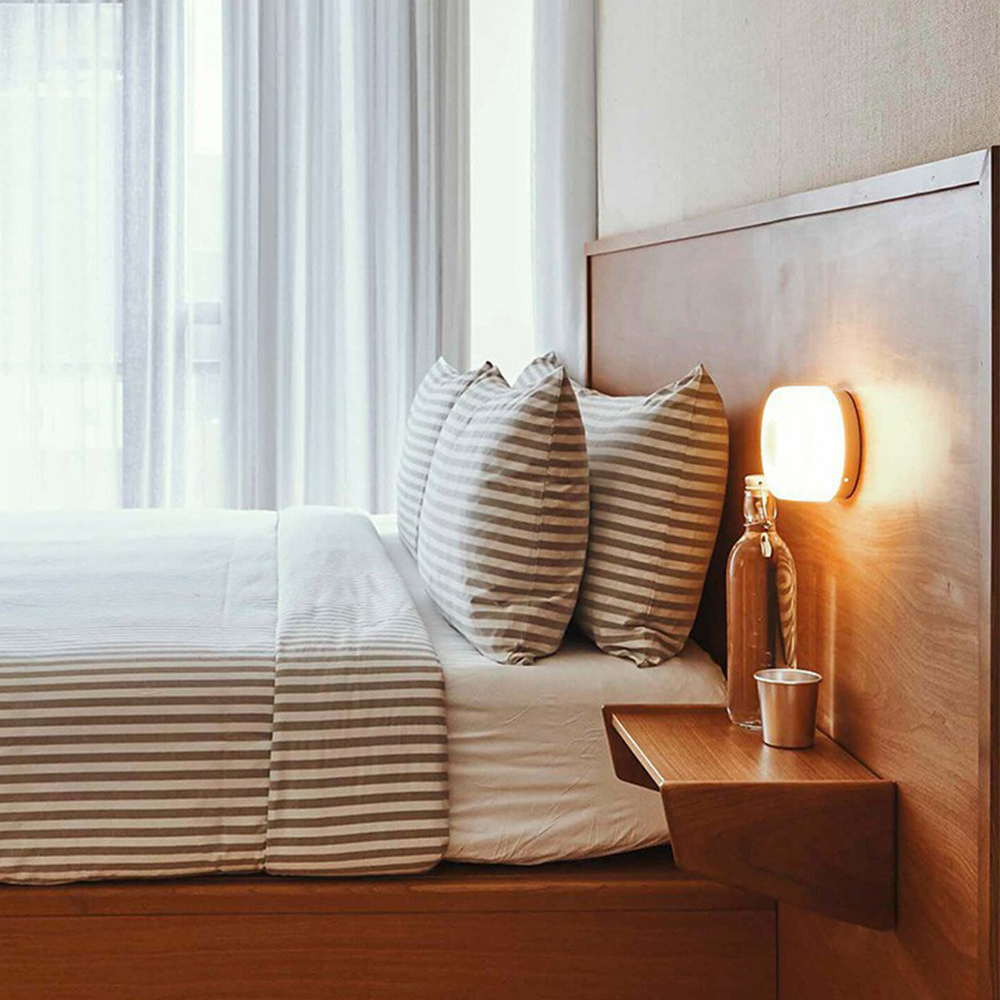 White round wall light beside striped bedding above a wooden shelf with a glass bottle and metal cup