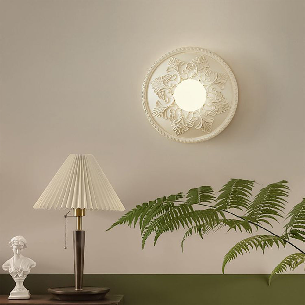 Ornate cream globe wall light above a pleated table lamp, bust and leafy plant on a two-tone wall