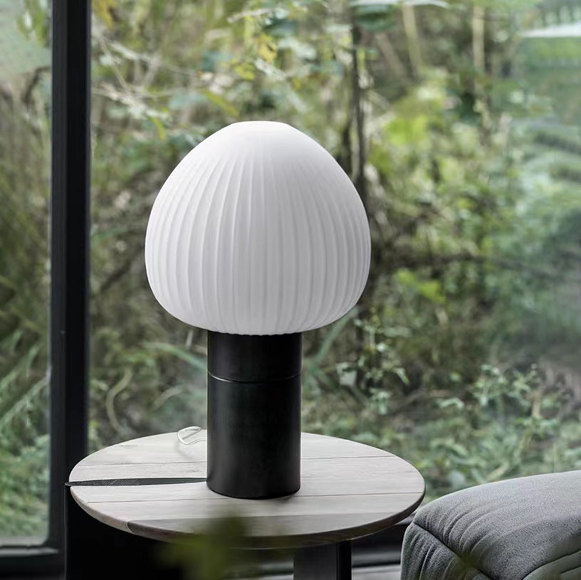 White ribbed-shade lamp on a slatted wooden side table beside black-framed windows, greenery and a grey sofa