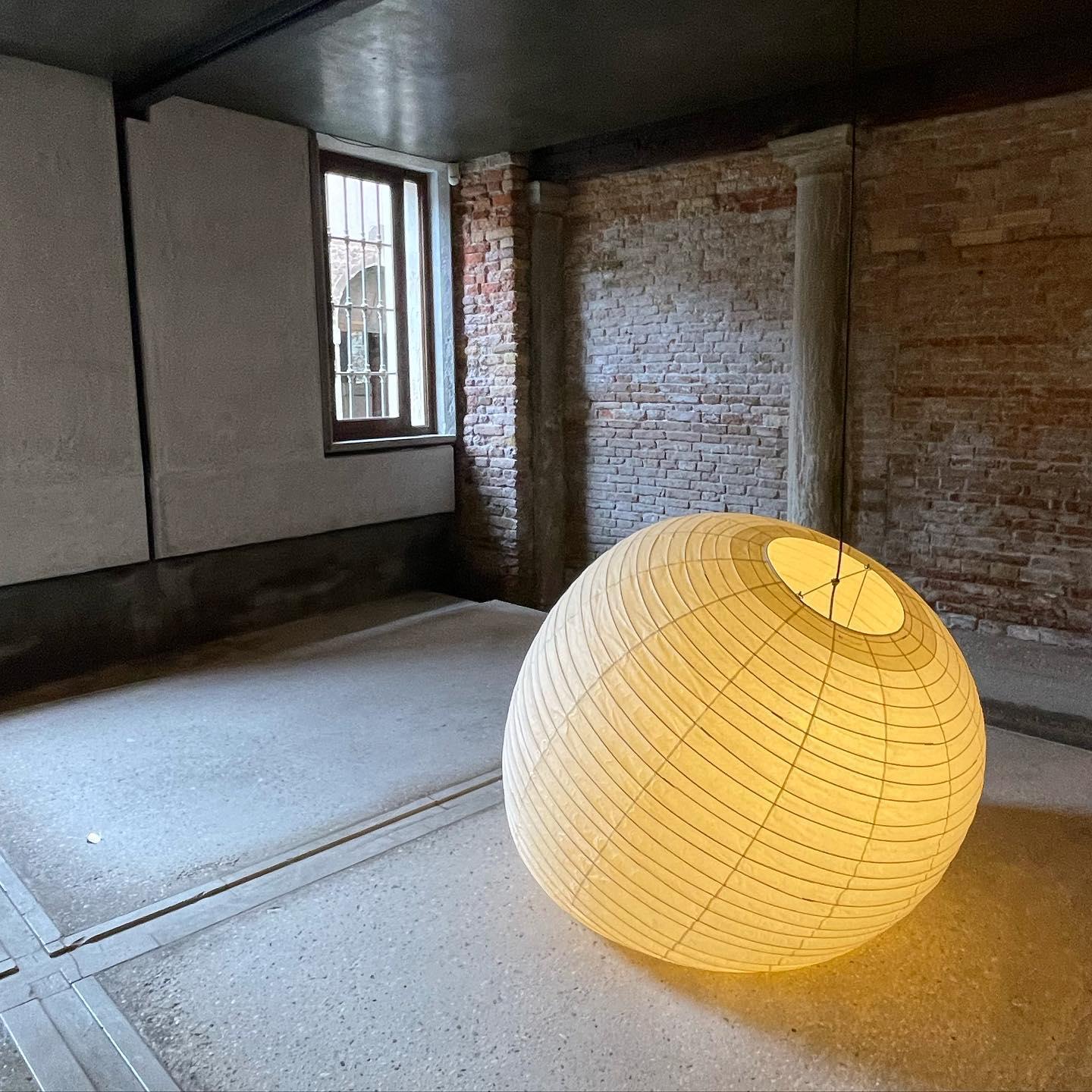 Tilted glowing paper globe with visible top opening and struts in a brick-walled gallery room