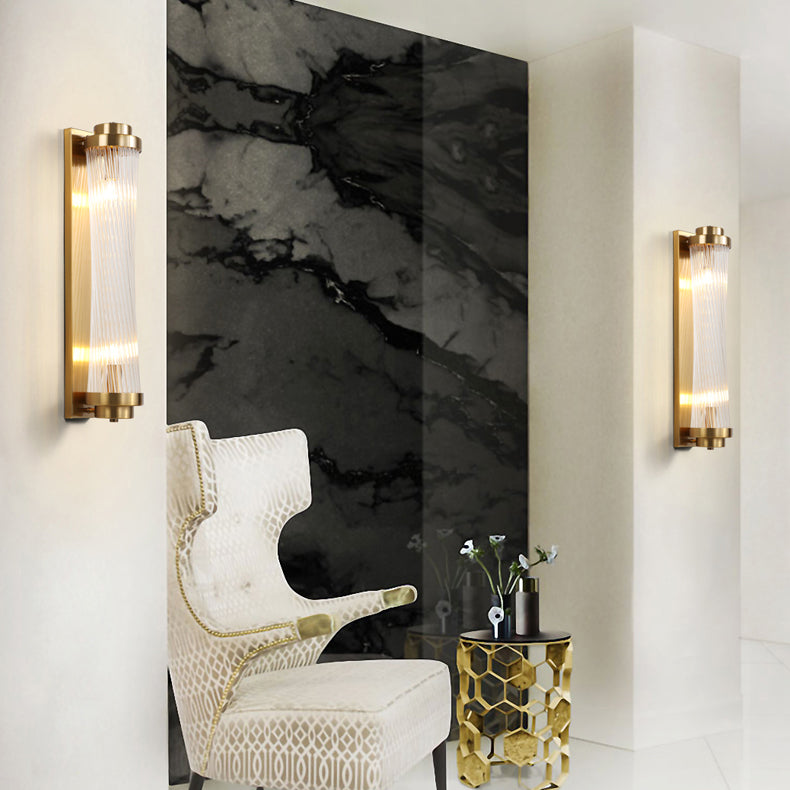 Two lit Gold rod wall lights around a dark veined wall panel, beside a cream patterned chair and gold geometric side table