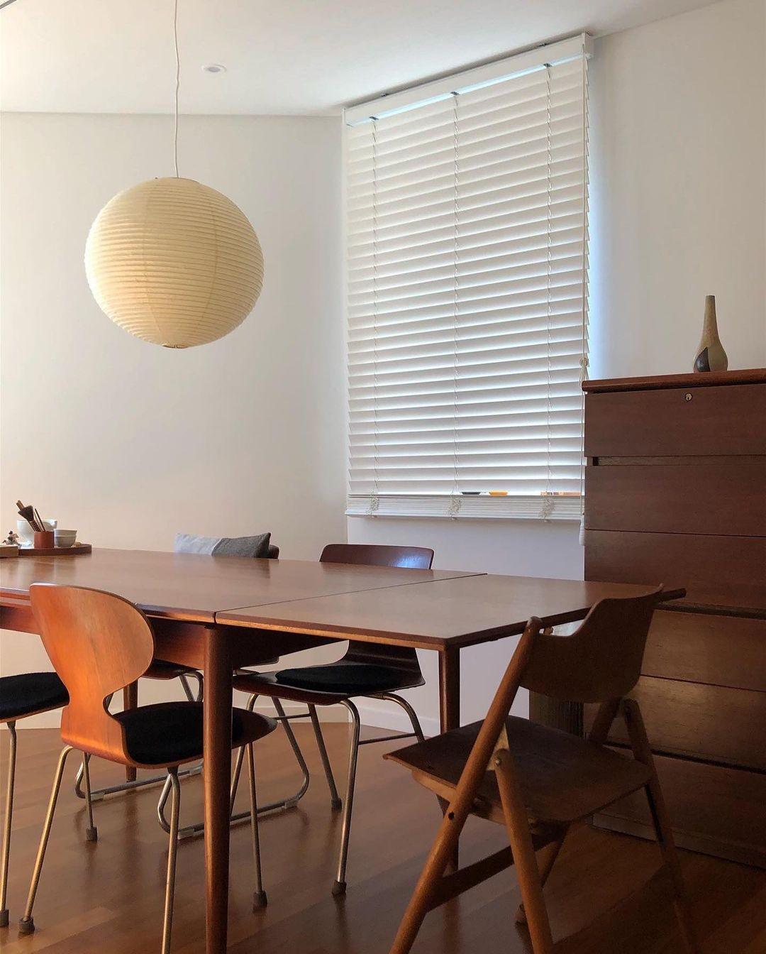 Paper globe above a rectangular wood dining table with mixed chairs before white blinds and a dresser
