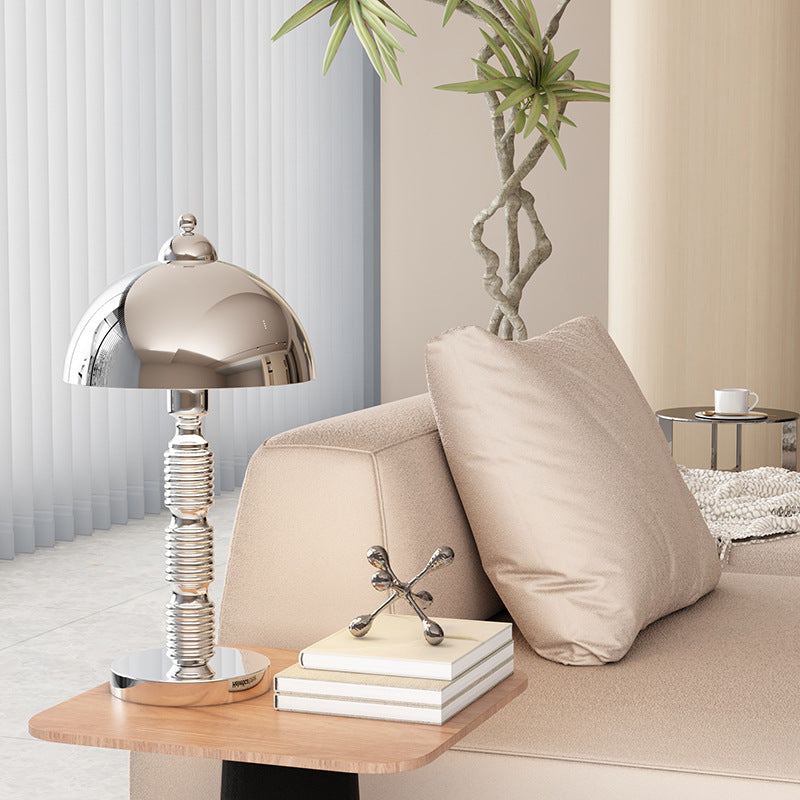 Chrome dome table lamp on a wooden sofa-side tray beside books and a sculptural ornament, with beige sofa and tall plant