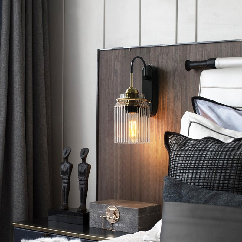 Ribbed glass wall sconce above a wood bedside panel with patterned pillows, two dark figures and a gray box nearby