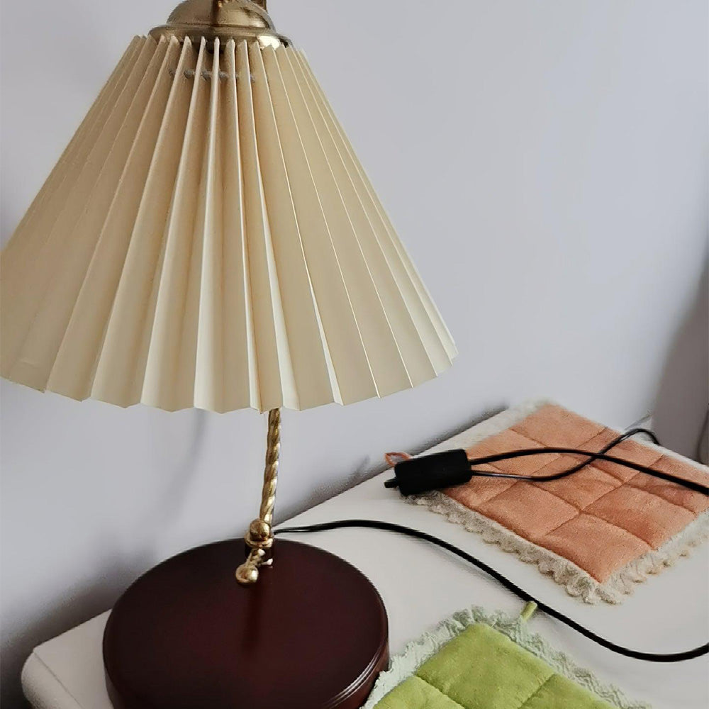 Beige pleated lamp on a white table beside patchwork cloth, with black cord and plug visible