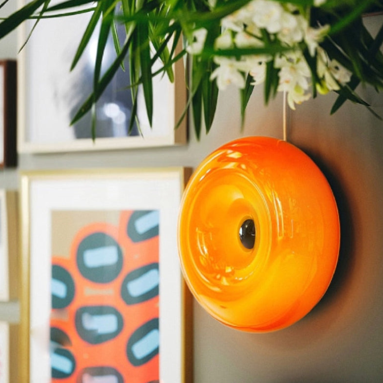 Close oblique orange donut wall light with reflective centre among framed prints and white flowers