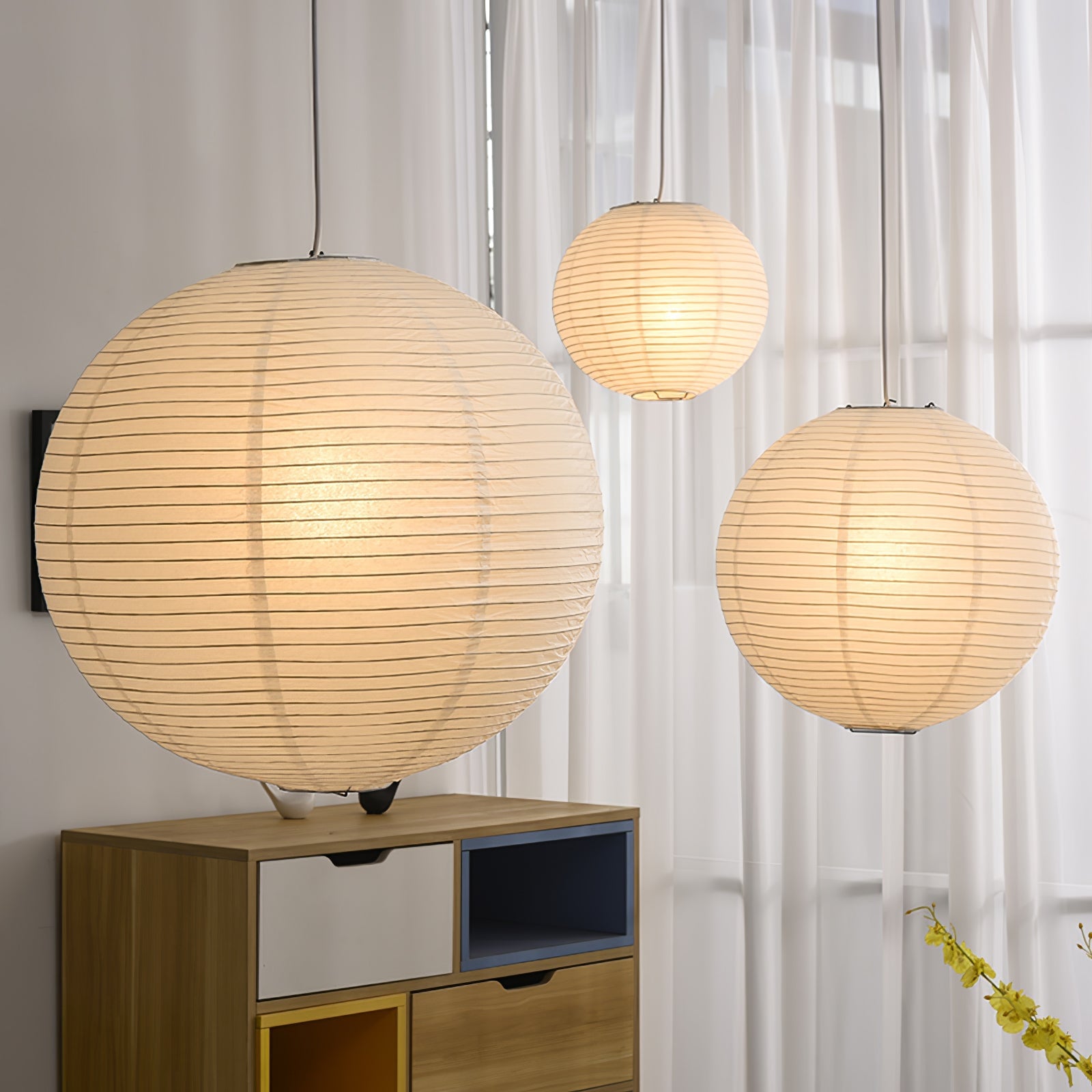 Three glowing paper globes at different heights over a colourful wood cabinet and sheer curtains