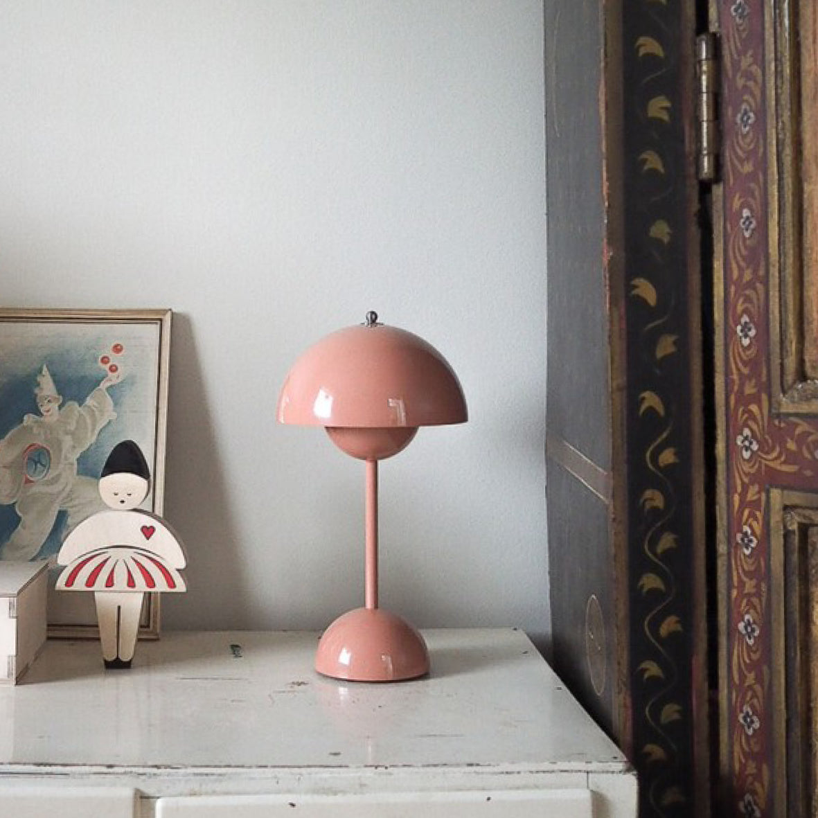 Pink dome lamp on a distressed white cabinet beside framed art and a decorated screen