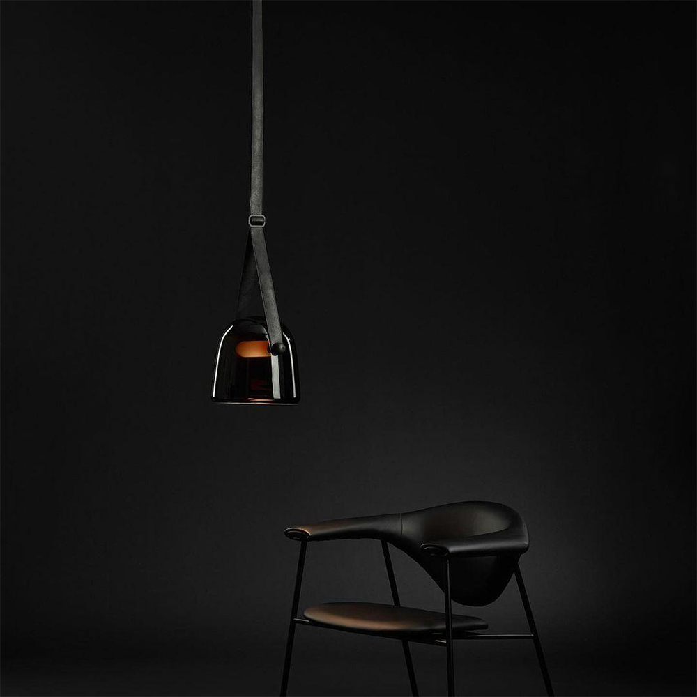 Black-strapped smoked glass pendant with warm-looking light bar against a dark background beside a black chair