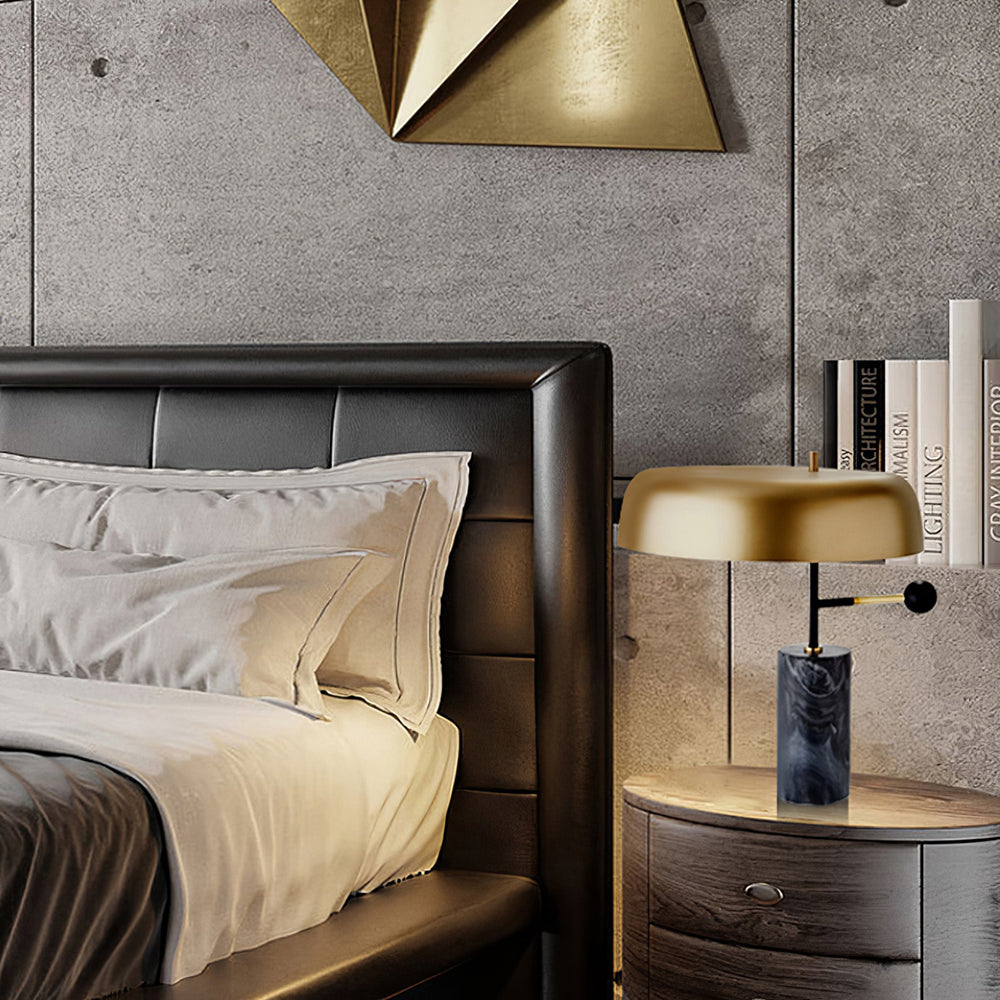 Gold-shade lamp on a round wood bedside cabinet beside dark headboard, pale pillows and upright books