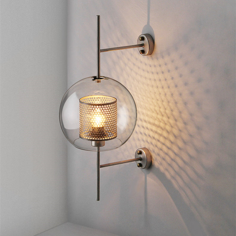 Clear globe wall sconce with perforated gold-tone inner cylinder and vertical rod against gray corner wall