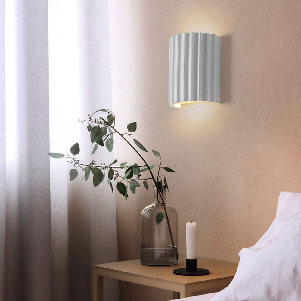 White fluted bedside wall light above a wooden table with branch vase and candle beside white curtains and bedding