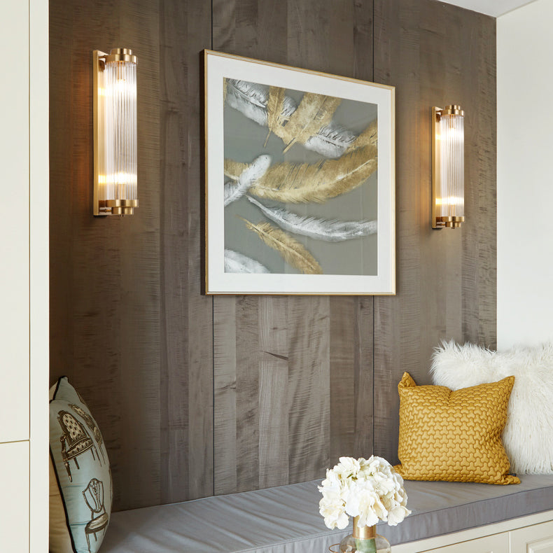 Two Gold Crystal Pillar wall lights flanking feather artwork above a cushioned bench on a wood wall