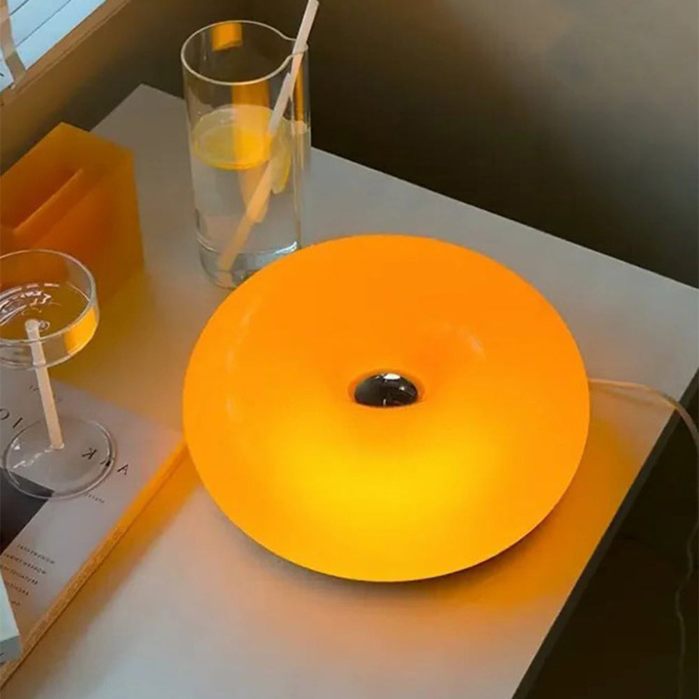 Overhead view of the glowing orange donut lamp on a pale table with a white cord, drinking glasses and a yellow box