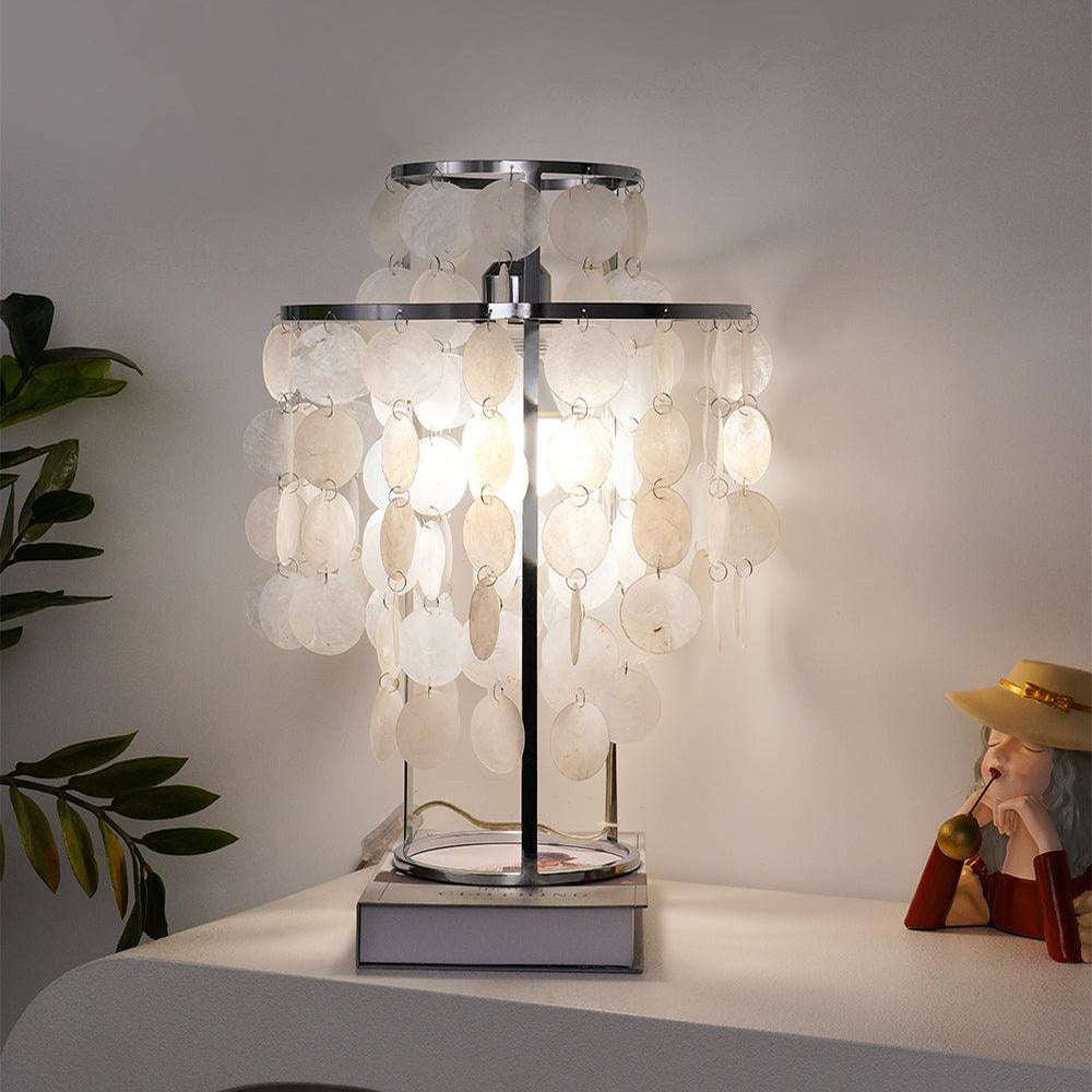 Lit silver-toned shell-disc lamp on a book and pale curved console, beside leafy plant and seated girl ornament