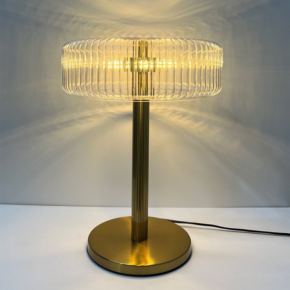 Illuminated ribbed-glass shade above a fluted gold-tone column and round brushed foot, with patterned wall light and a black cord to the right