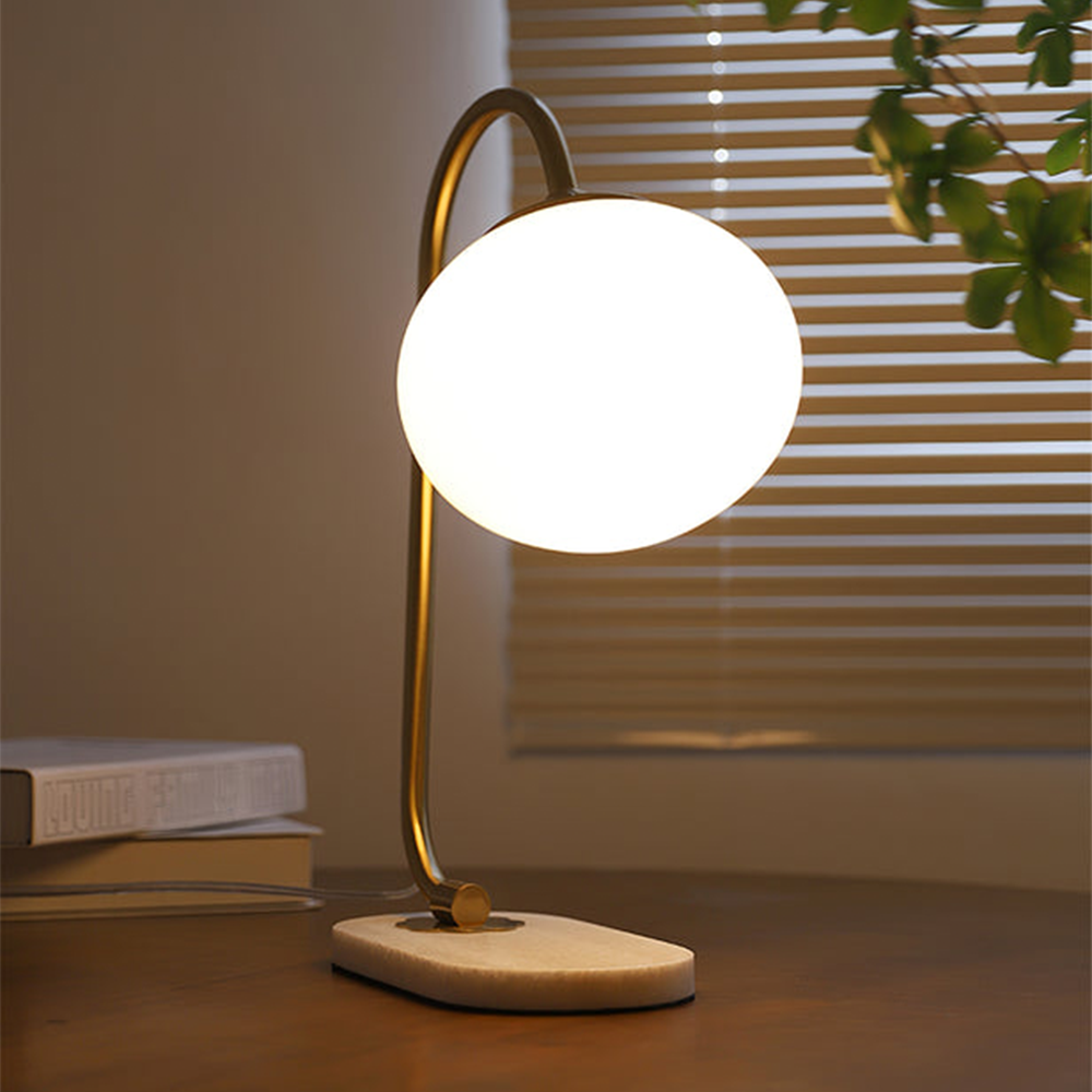 Glowing white globe lamp on a wood table beside books and window blinds, with a cord behind