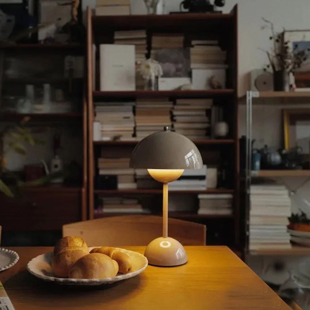Pale grey lamp with a lit lower bowl on a wooden table beside bread and bookcases