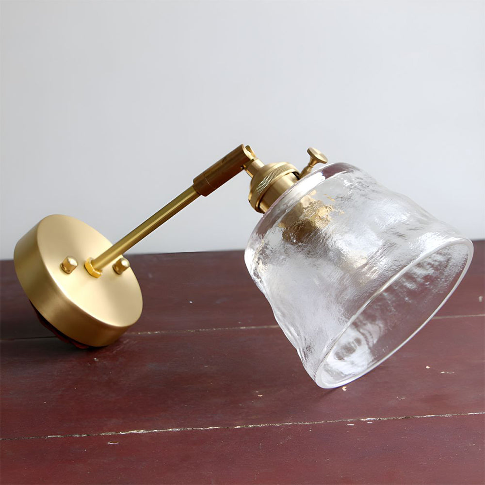 Clear bell shade with open rim, short metal arm and round wallplate lying on reddish wooden tabletop