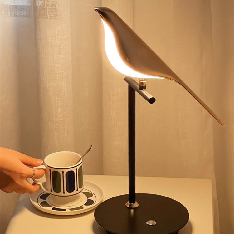 Single glowing gold-tone bird lamp on a pale table beside a cup and saucer against cream curtains