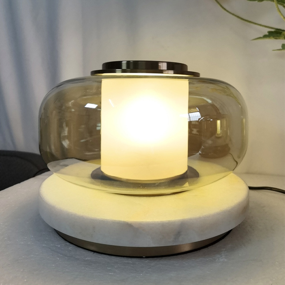 Light-brown glass lamp with a glowing pale cylinder and light circular base near pale curtains, with a cord extending right