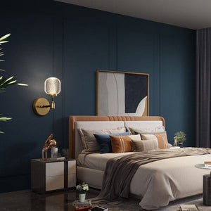Gold wall light beside a pale upholstered bed against teal panelling and gold-framed artwork