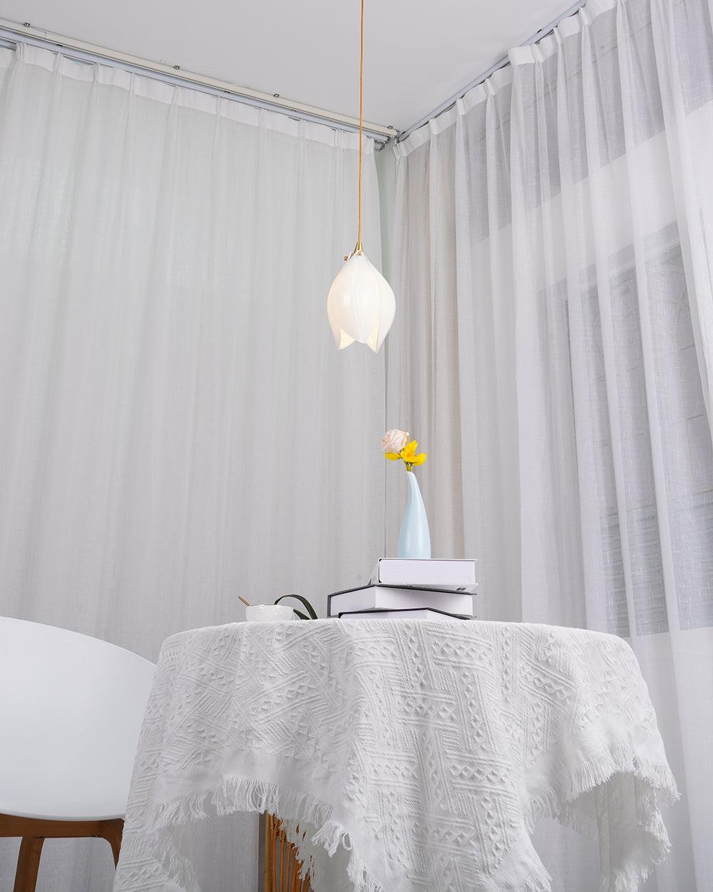 Single flower pendant above a cloth-covered table and books near sheer curtains