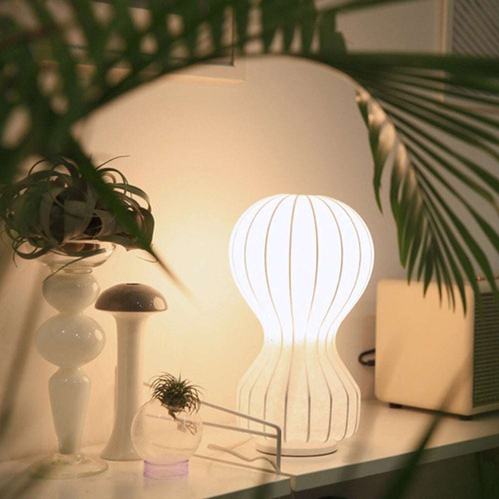 Glowing ribbed lamp with a round foot on a white surface among sculptural vases, plants and a separate mushroom-shaped lamp