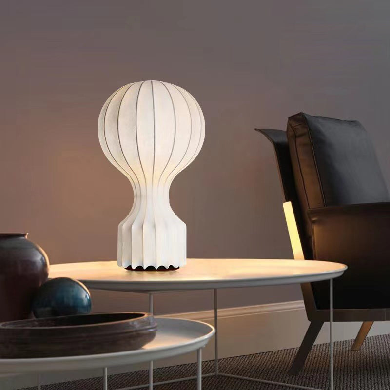 Wide view of the lit ribbed lamp on a pale round table beside a dark wooden chair and foreground ceramic vessels