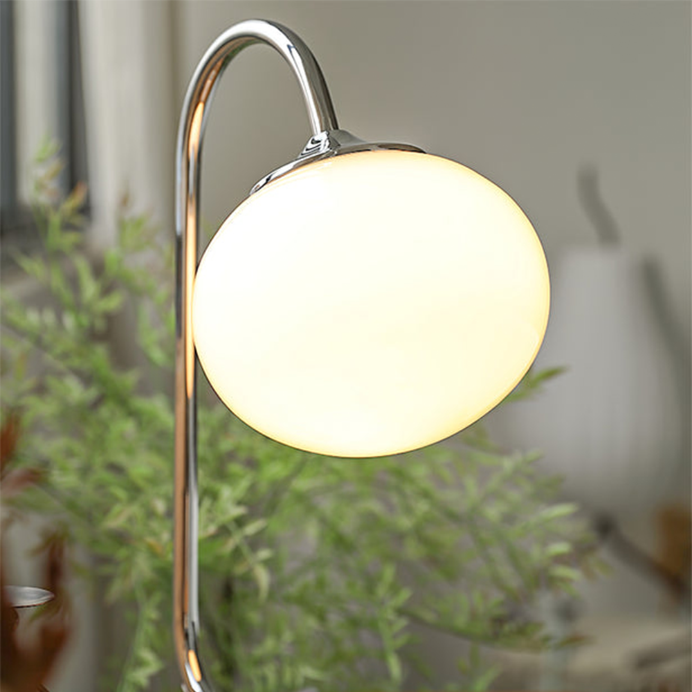 Illuminated pale globe and chrome-finish curved neck with green plant leaves and curtains behind