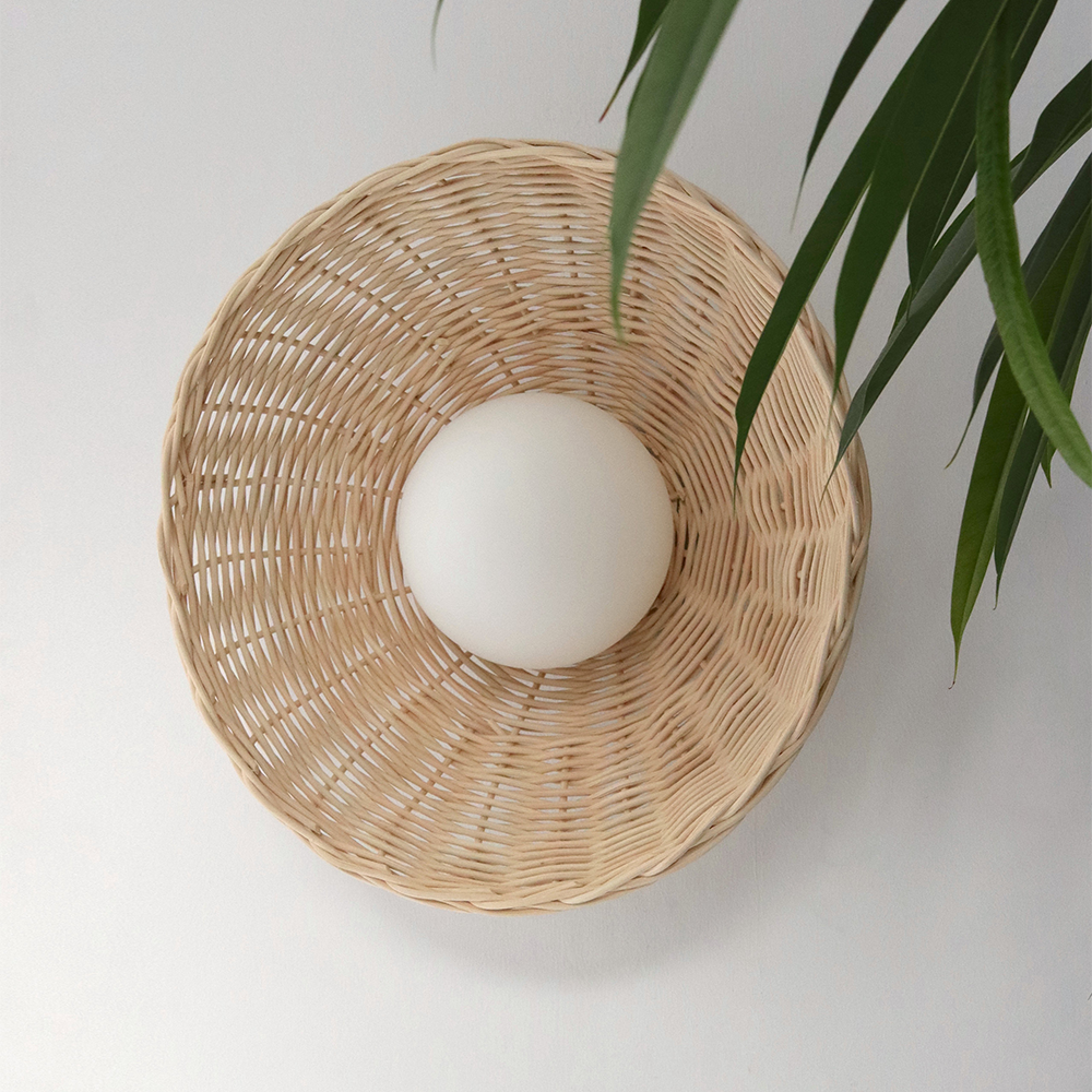Unlit pale woven round shade and white matte globe on a light wall beside green leaves