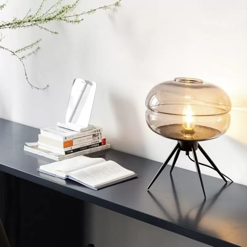 Lit rounded glass lamp on black splayed legs beside an open book, stacked books and a propped phone on a dark desk