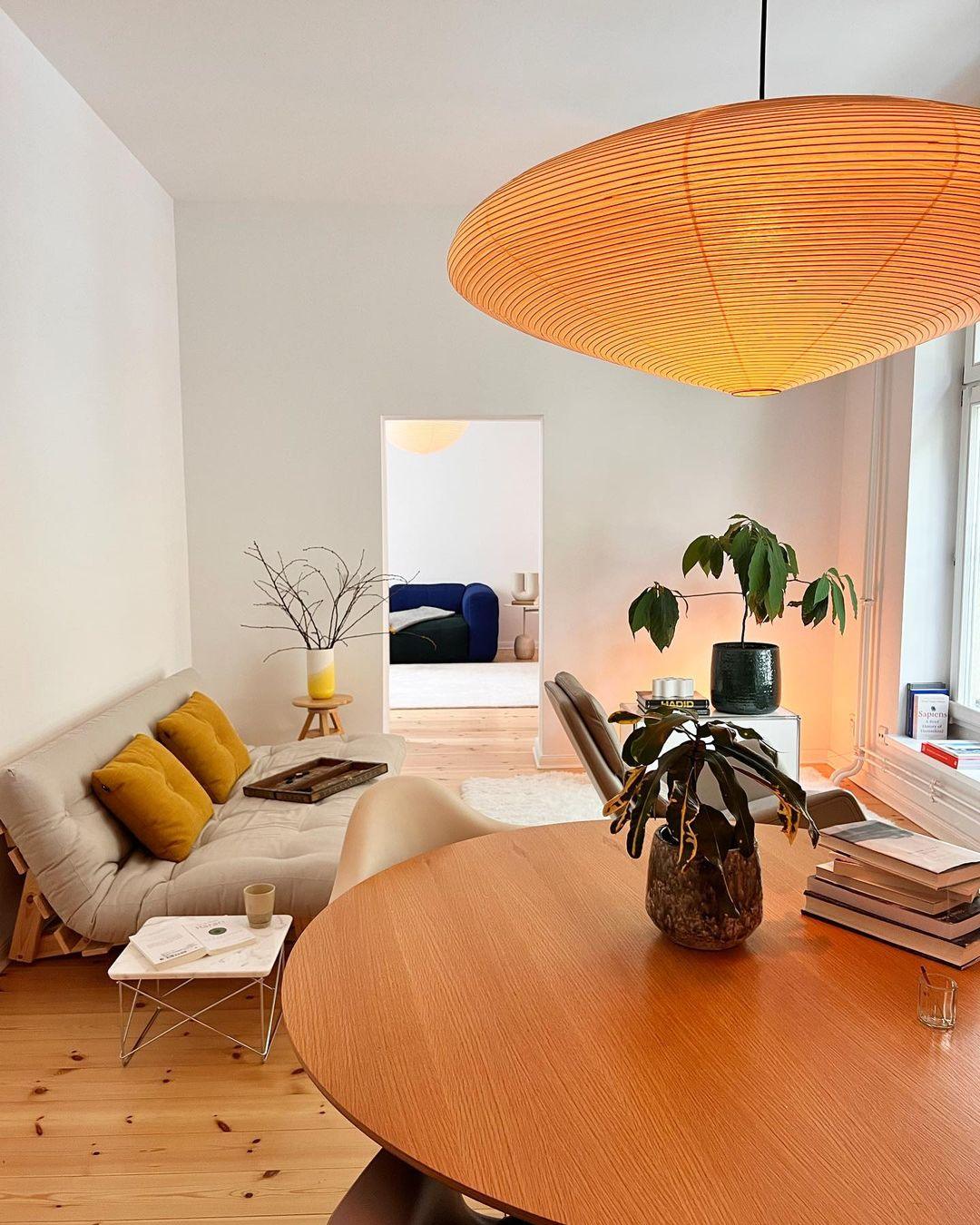 Orange-looking glow from a flattened shade above a round wood table beside a pale sofa with yellow cushions