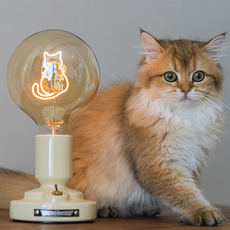 Pale-base cat-outline globe lamp with small metal lever and front badge beside a live cat on a wood surface