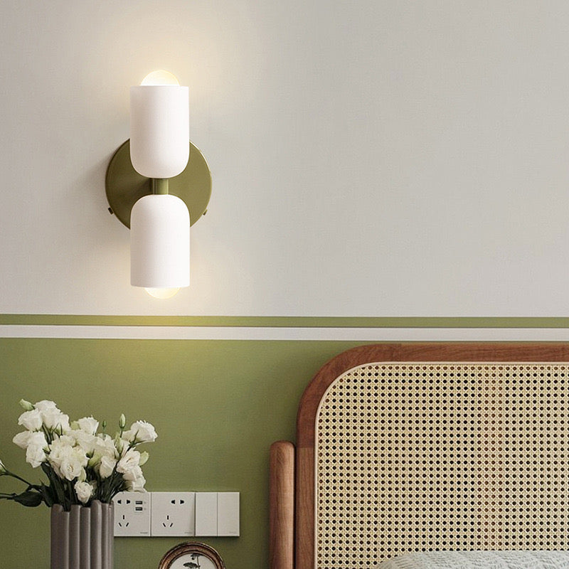 Lit green double-head sconce on a two-tone bedroom wall beside a cane headboard and flowers in a metal vase