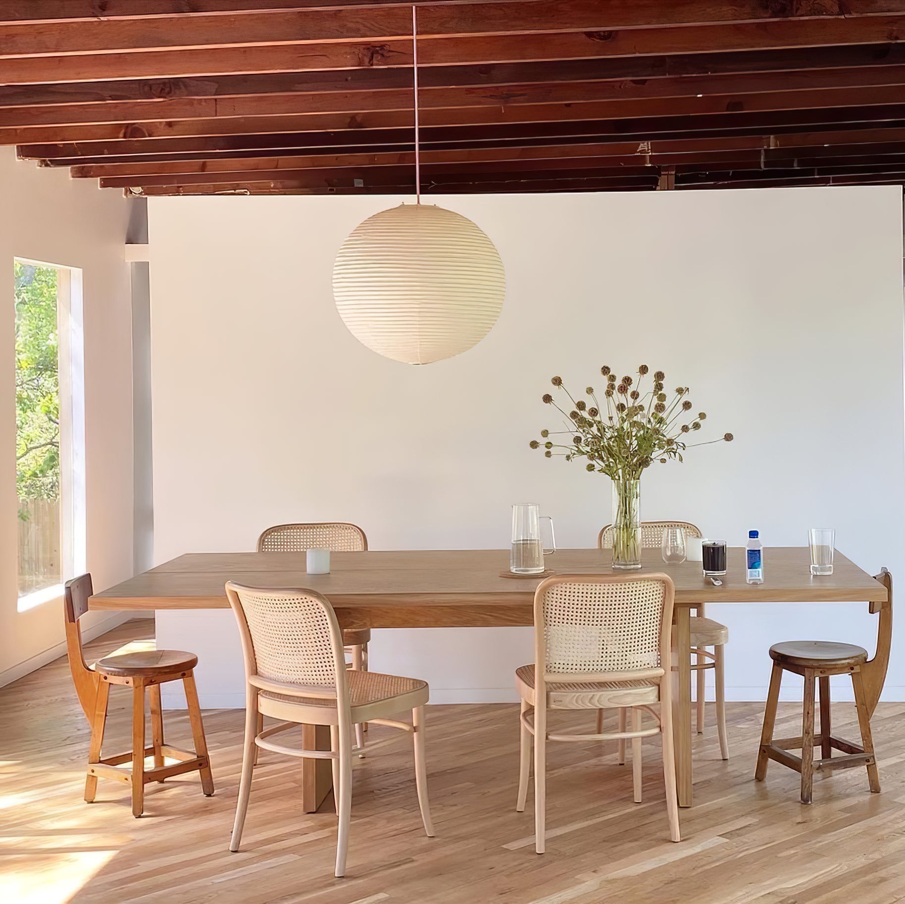Paper globe over a wood dining table with cane-backed chairs, stools and flower vase beneath timber beams