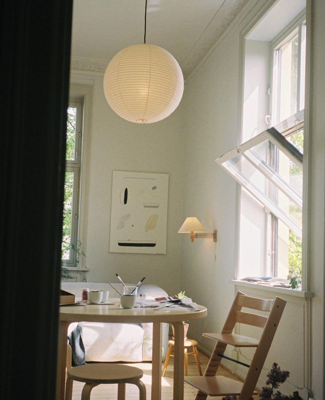 Small glowing paper globe above a cream round desk with cups and brushes beside a folding wood chair