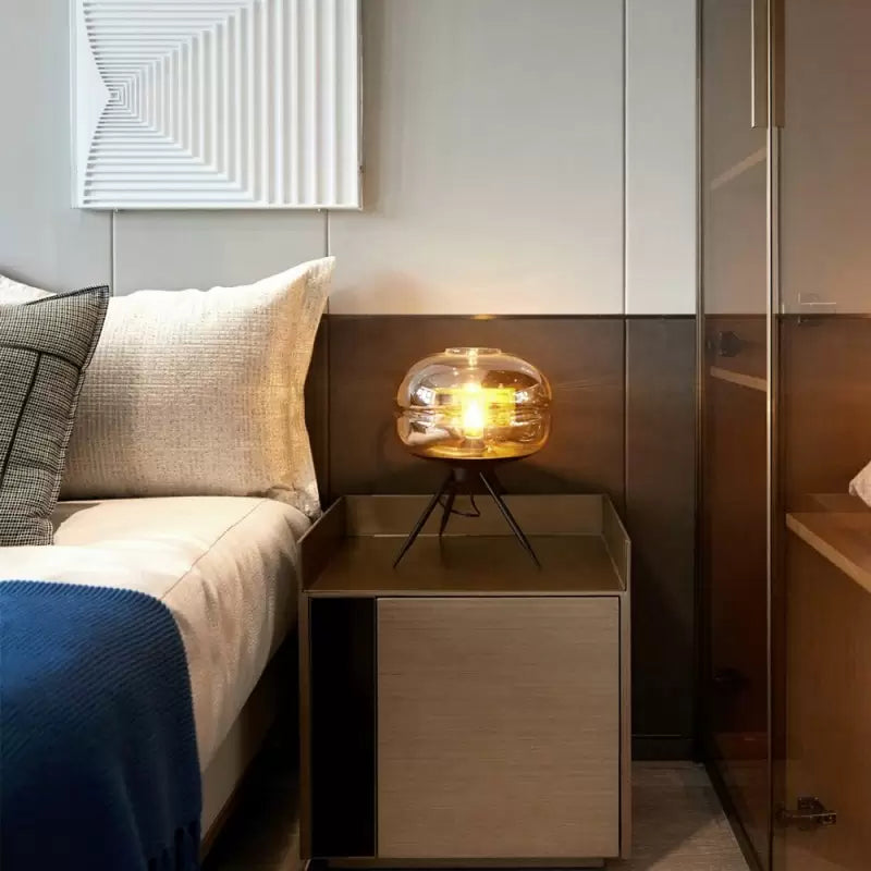 Glowing light-brown glass lamp on a tray-top bedside cabinet beside a bed with pale cushions and a blue cover